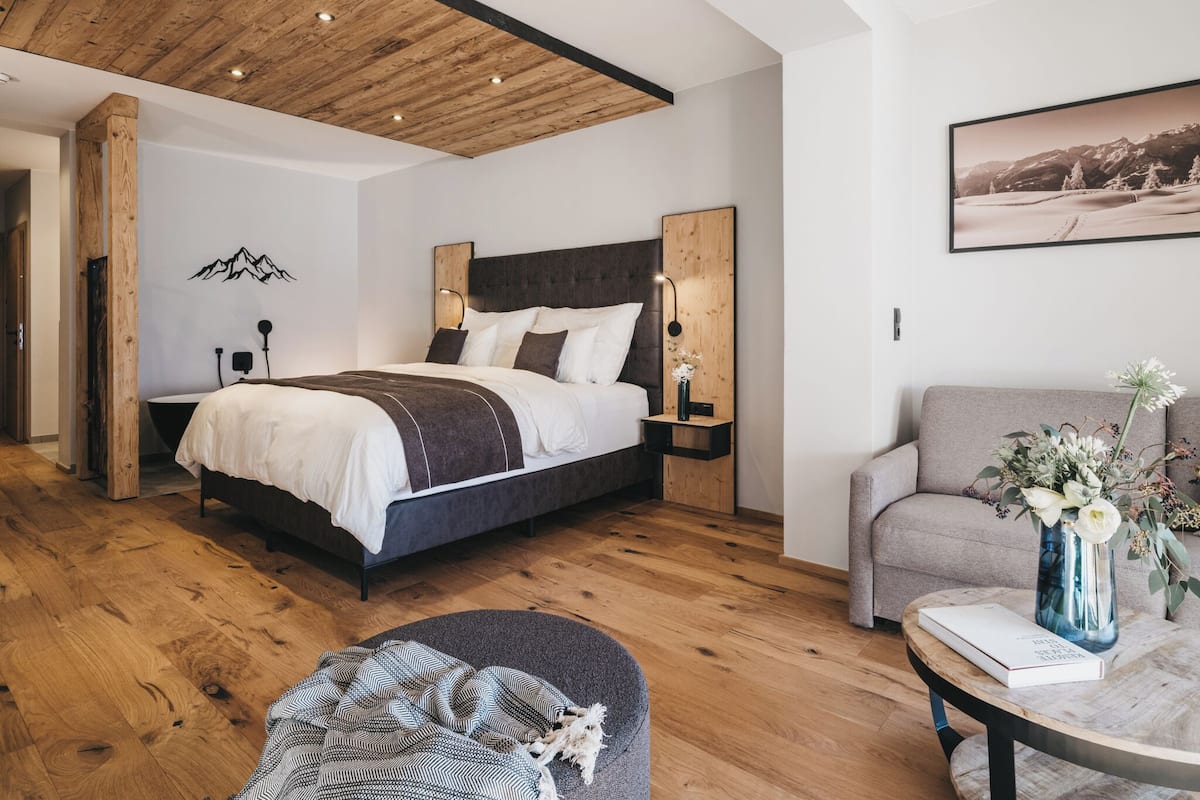 Un dormitorio moderno con techo de madera, una cama grande, un sofá gris y una mesa de centro redonda con un libro y flores.