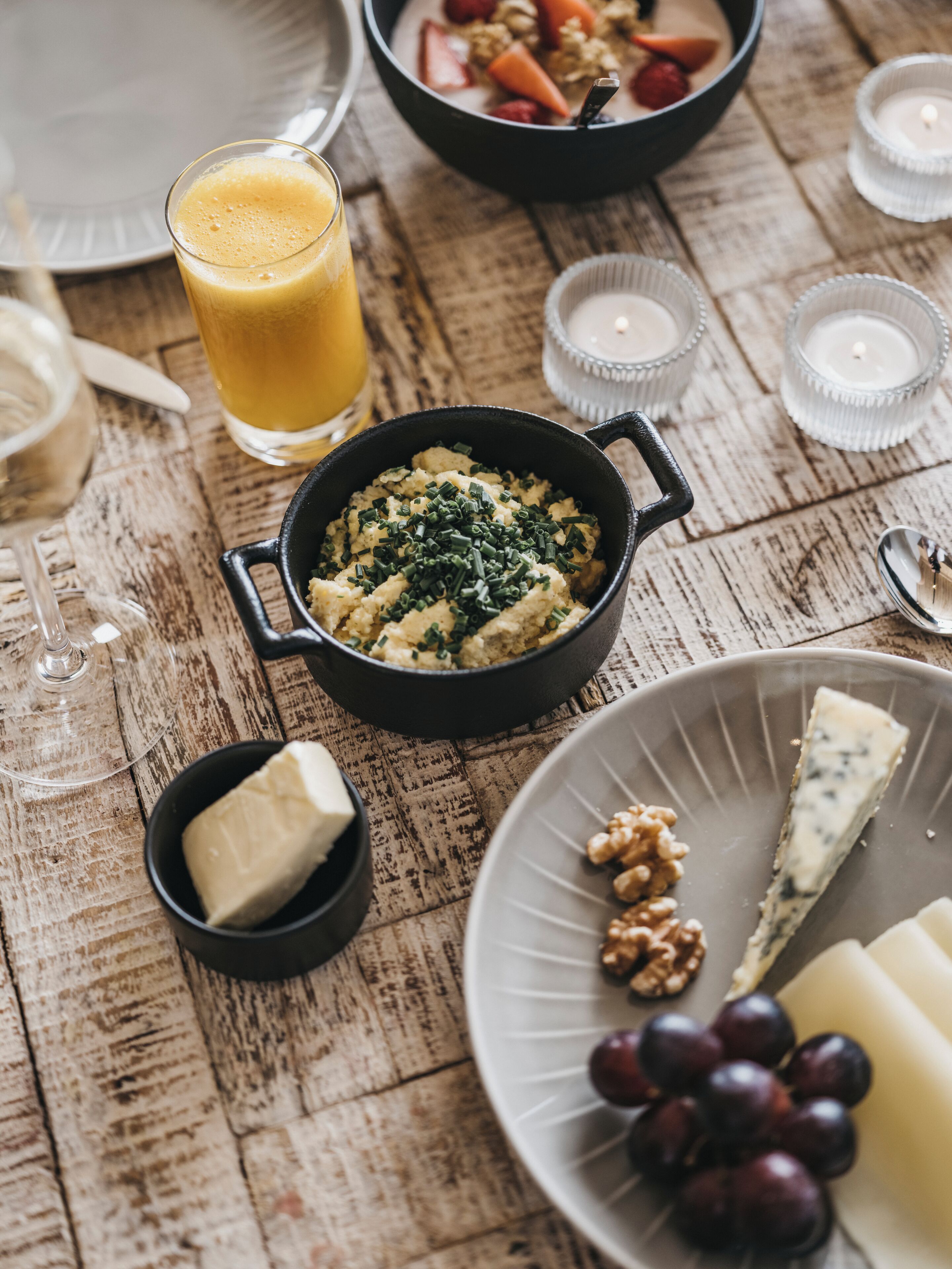 Ein Holztisch mit verschiedenen Speisen, darunter eine Schüssel Kartoffelpüree, ein Teller mit Käse und Trauben, ein Glas Orangensaft und eine Schüssel Obstsalat.