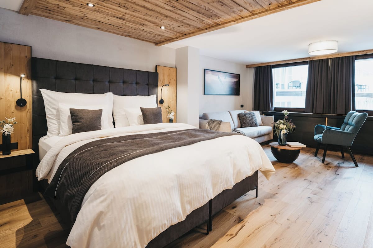 Una habitación de hotel moderna con techo de madera, una cama grande con ropa de cama blanca y marrón, un sofá, una silla y un ventanal con cortinas.