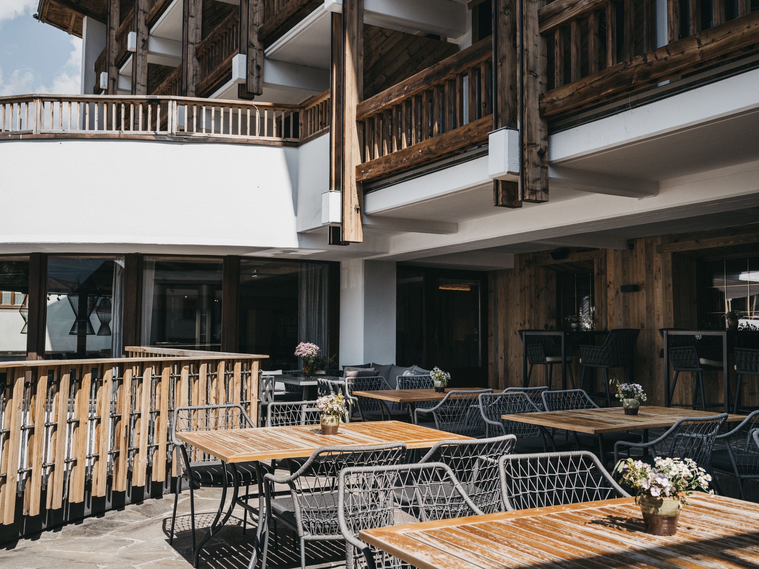 An outdoor seating area with wooden tables and chairs, a balcony with wooden railings, and a view of the sky.