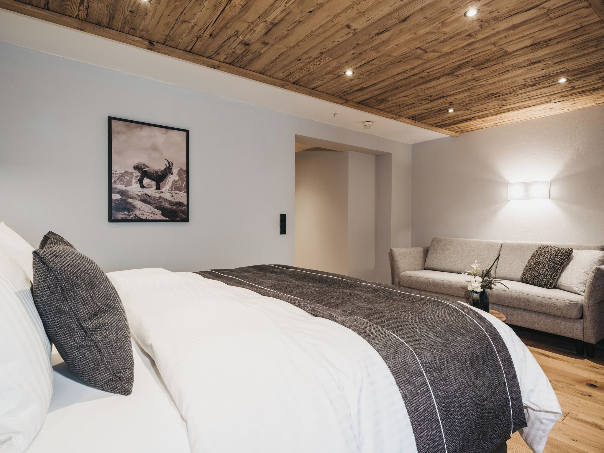 A bedroom with a wooden ceiling, a bed with white and dark bedding, a gray sofa, and a framed picture on the wall.
