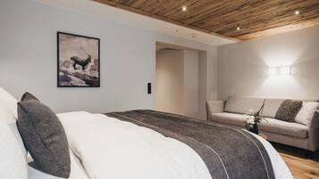 Ein Schlafzimmer mit Holzbalkendecke, einem Bett mit weißer und dunkler Bettwäsche, einem grauen Sofa und einem gerahmten Bild an der Wand.