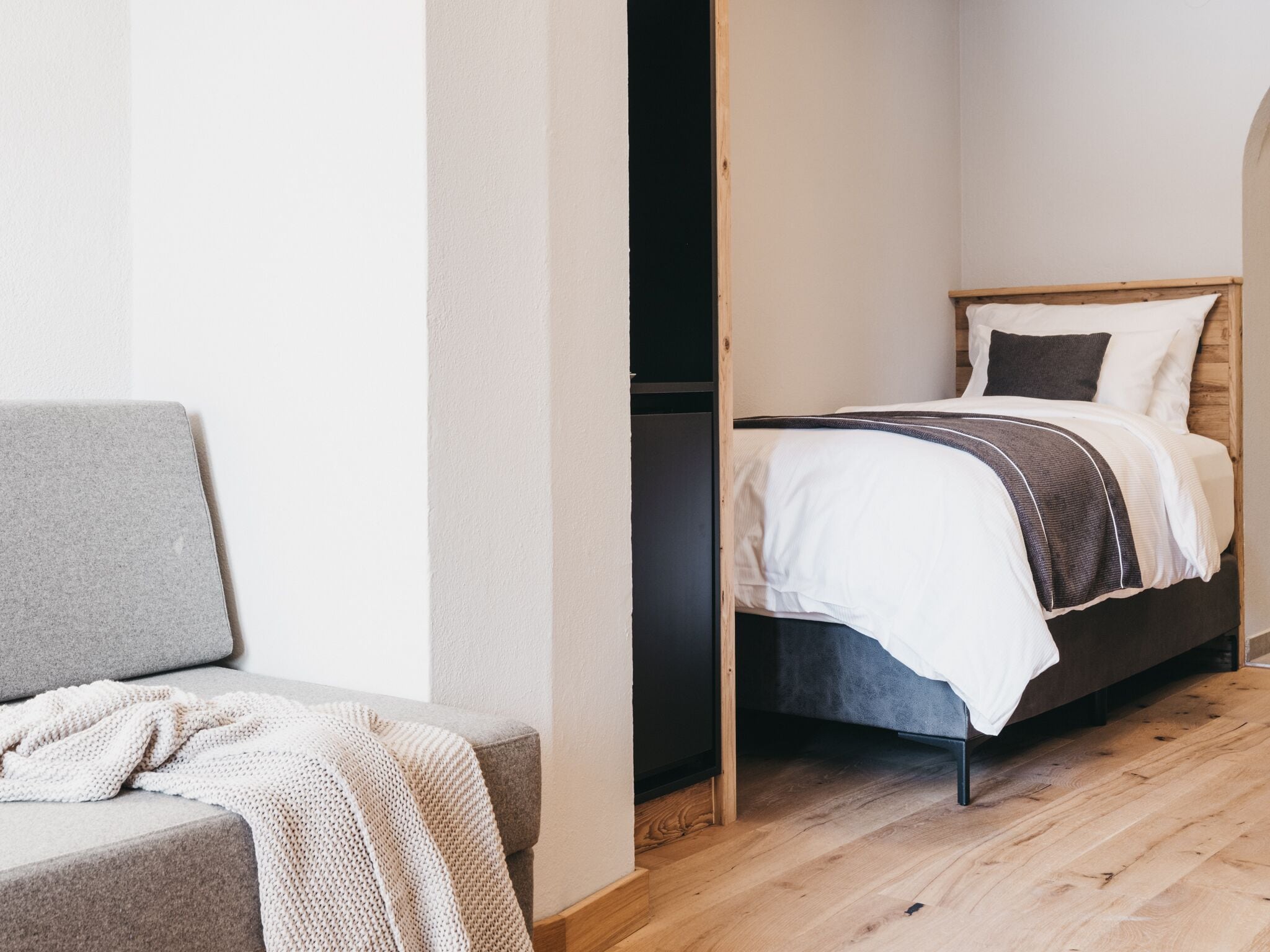 A modern bedroom with a bed, a grey sofa, and a wooden floor.