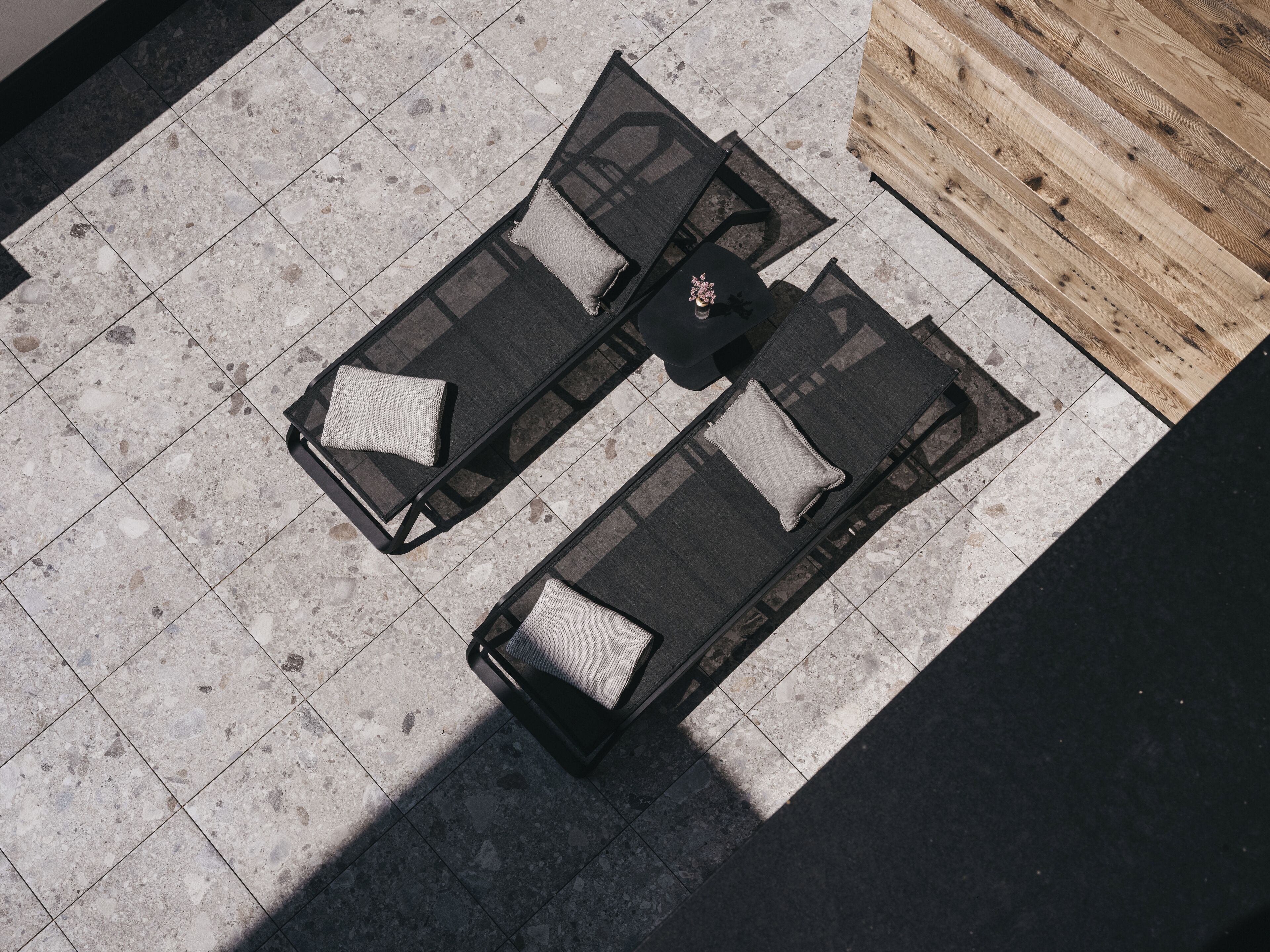 An outdoor seating area with two black lounge chairs and a small table, set on a tiled floor with a wooden partition in the background.