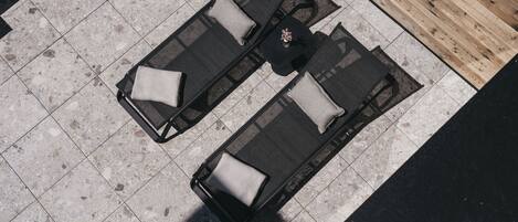 An outdoor seating area with two black lounge chairs and a small table, set on a tiled floor with a wooden partition in the background.