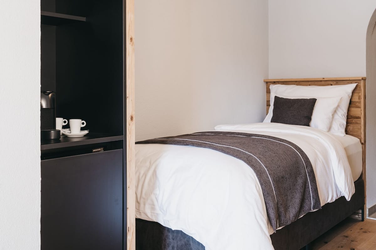 Una cama individual con cabecera de madera, ropa blanca de cama y una manta de color marrón oscuro. Una cafetera y tazas están sobre una repisa negra a la izquierda.