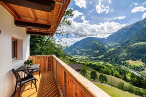 A wooden balcony with a table and chairs, overlooking a valley with mountains and a river.
