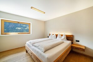 A bedroom with a large window, a bed with white linens, a wooden headboard, and a bedside table with a drawer.