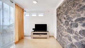 A room with a stone wall, a flat-screen TV on a wooden TV stand, a sliding glass door with orange curtains, and a tiled floor. - Holiday Home 'Simão House' with Private Terrace, Private Garden and Wi-Fi (Calheta)
