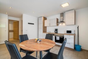 Une cuisine moderne avec des meubles blancs, un four à micro-ondes et un four traditionnel. On y trouve une table à manger avec des chaises et un saladier rempli de fruits.