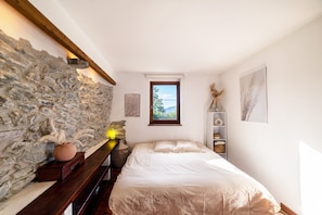 A bedroom with a stone wall, a bed, a window, a shelf with shoes, and a vase with dried plants.