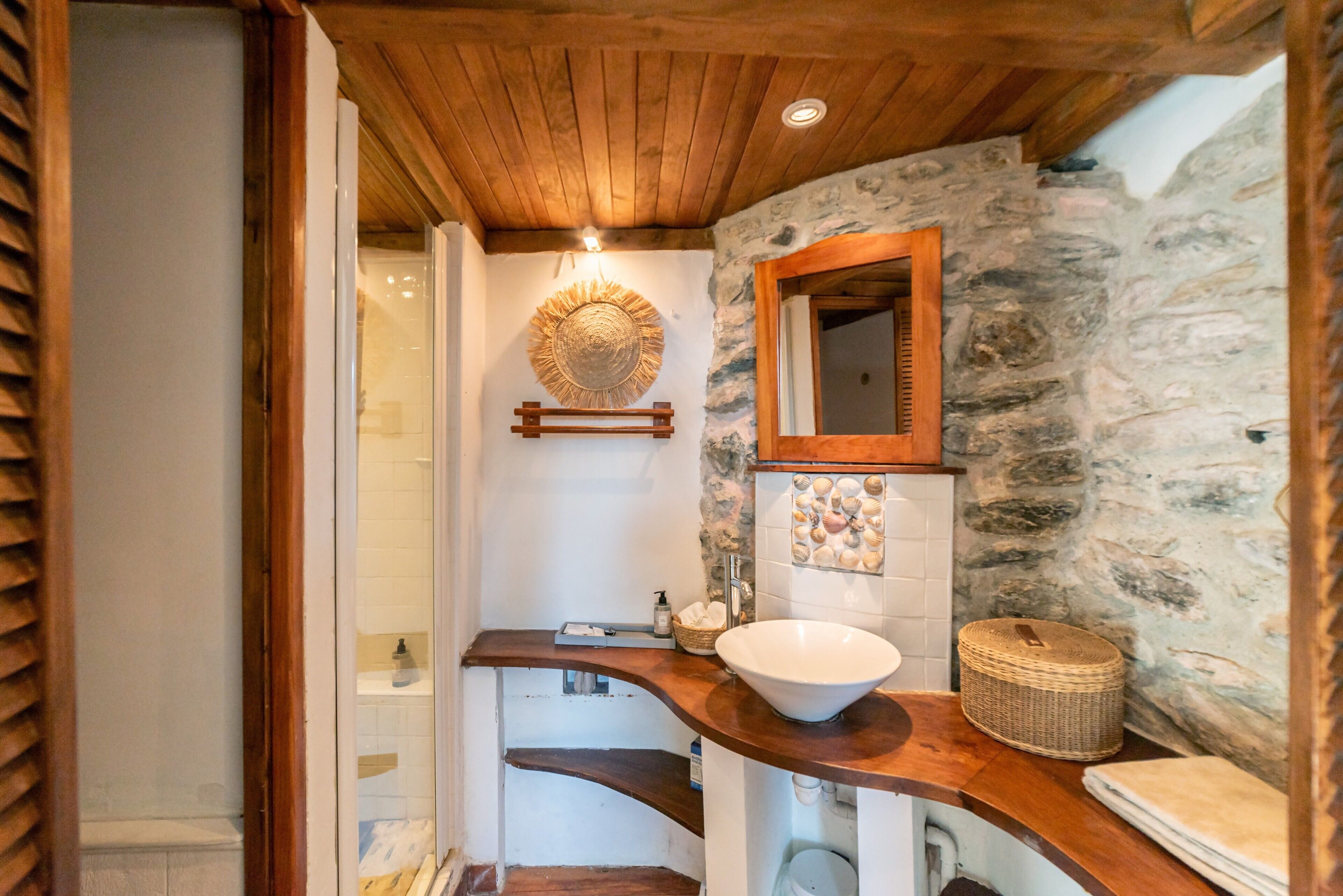 Une salle de bain avec un mur en pierre, un plafond en bois, un grand miroir, un lavabo blanc et une étagère en bois.