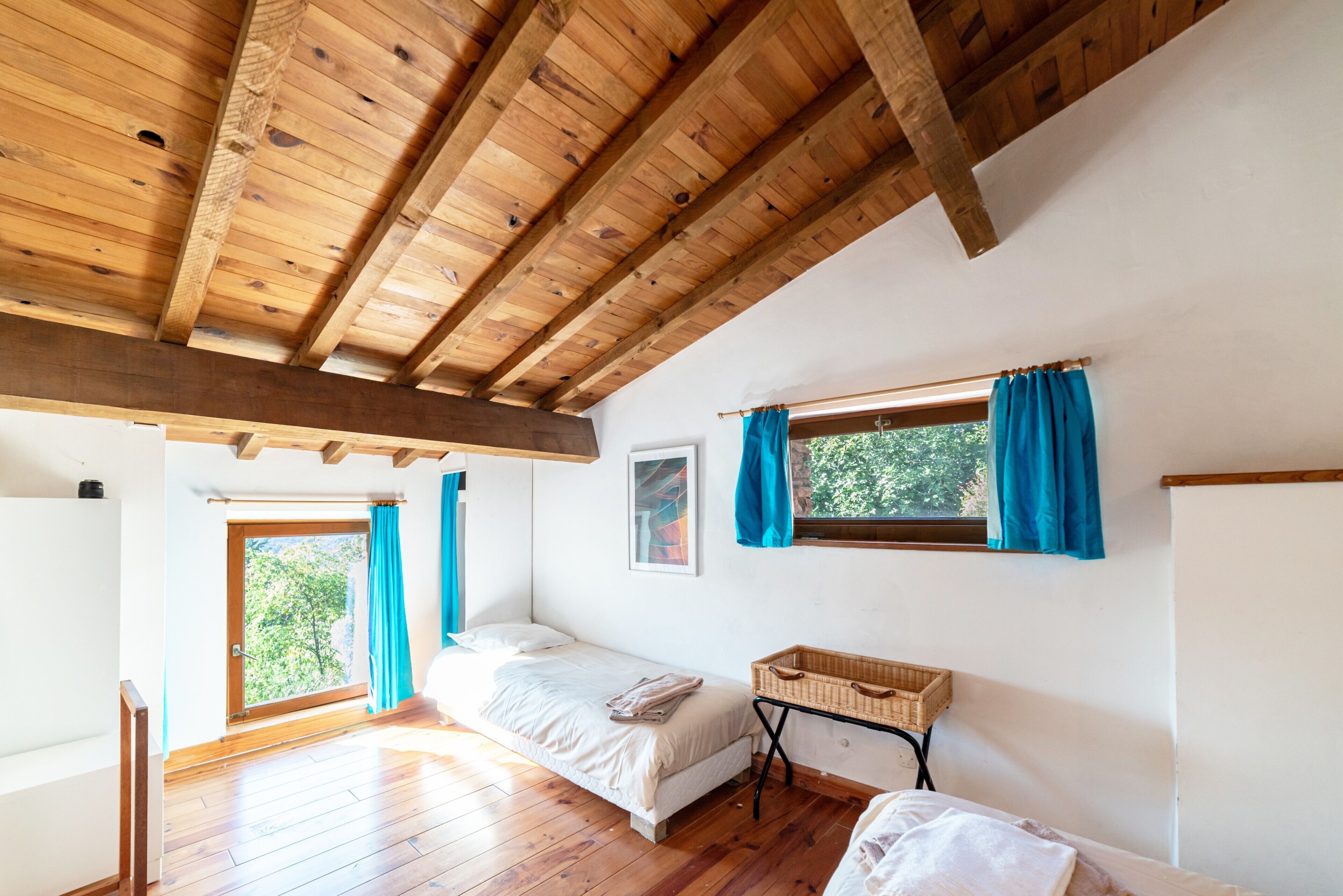 A room with wooden beams, a bed, a window with curtains, a small table, and a door leading outside.