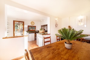A dining area with a wooden table, chairs, a potted plant, and a kitchen in the background.