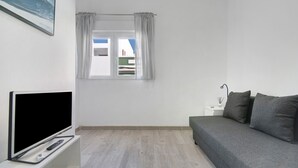 Ett rum med en platt-TV på ett stativ, en grå soffa, ett litet sängbord med en lampa och ett fönster med gardiner.