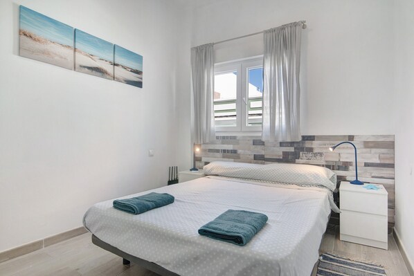 A bedroom with a bed, a nightstand, a lamp, a window with curtains, and a wall-mounted artwork.