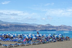 A beach with rows of sun loungers and umbrellas, a clear sky, and mountains in the background.