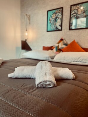 A neatly made bed with a rolled towel in the center, a patterned brown bedspread, and two abstract paintings on the wall.