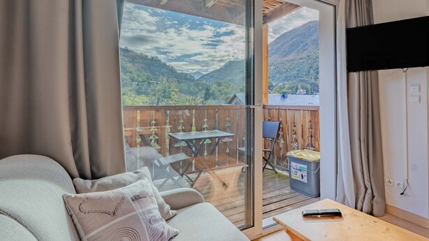 Un balcon avec vue sur les montagnes, un canapé avec des coussins, une table en bois et une télécommande.