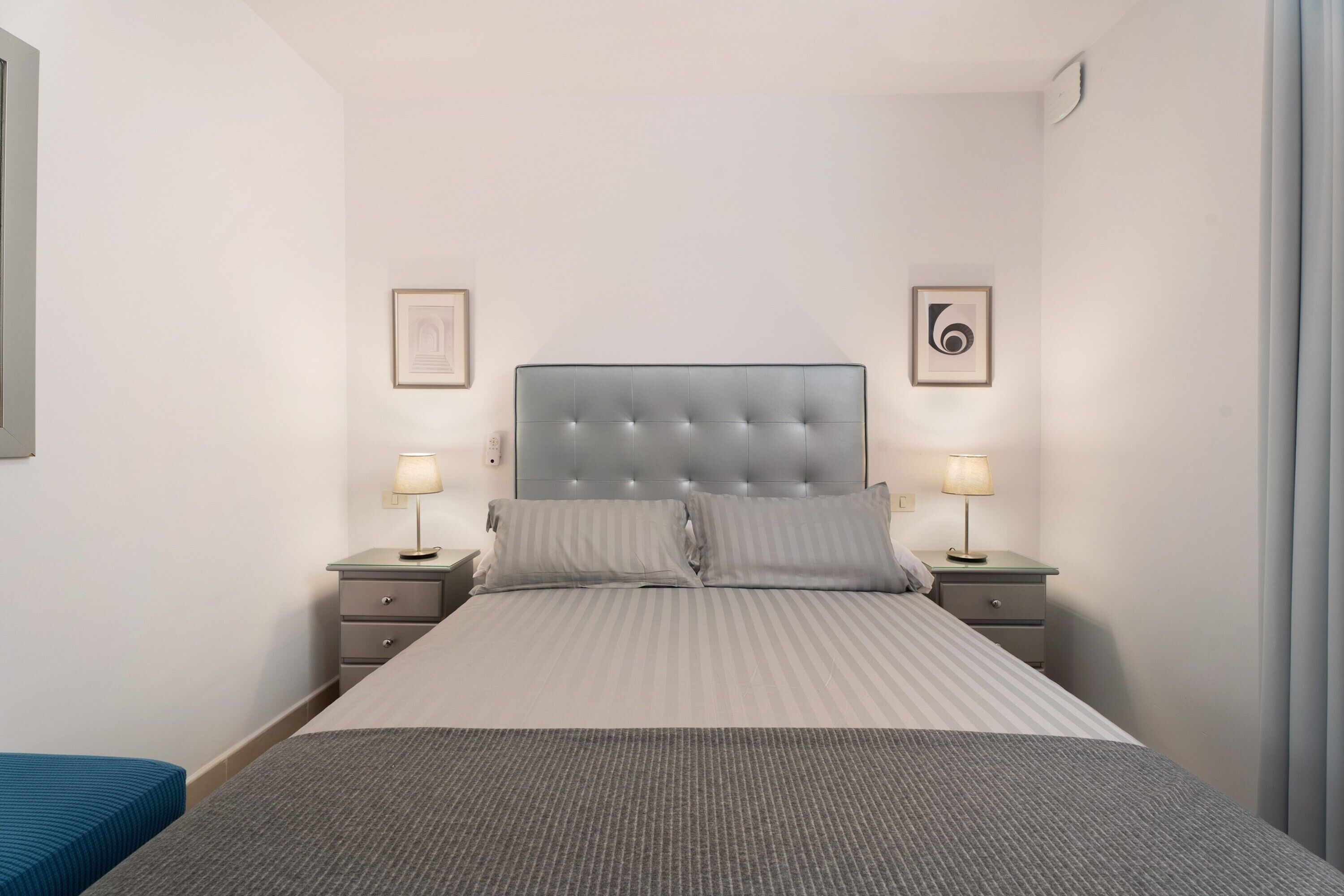 A neatly arranged hotel room with a large bed, two bedside tables with lamps, a small blue bench, and framed artwork on the walls.