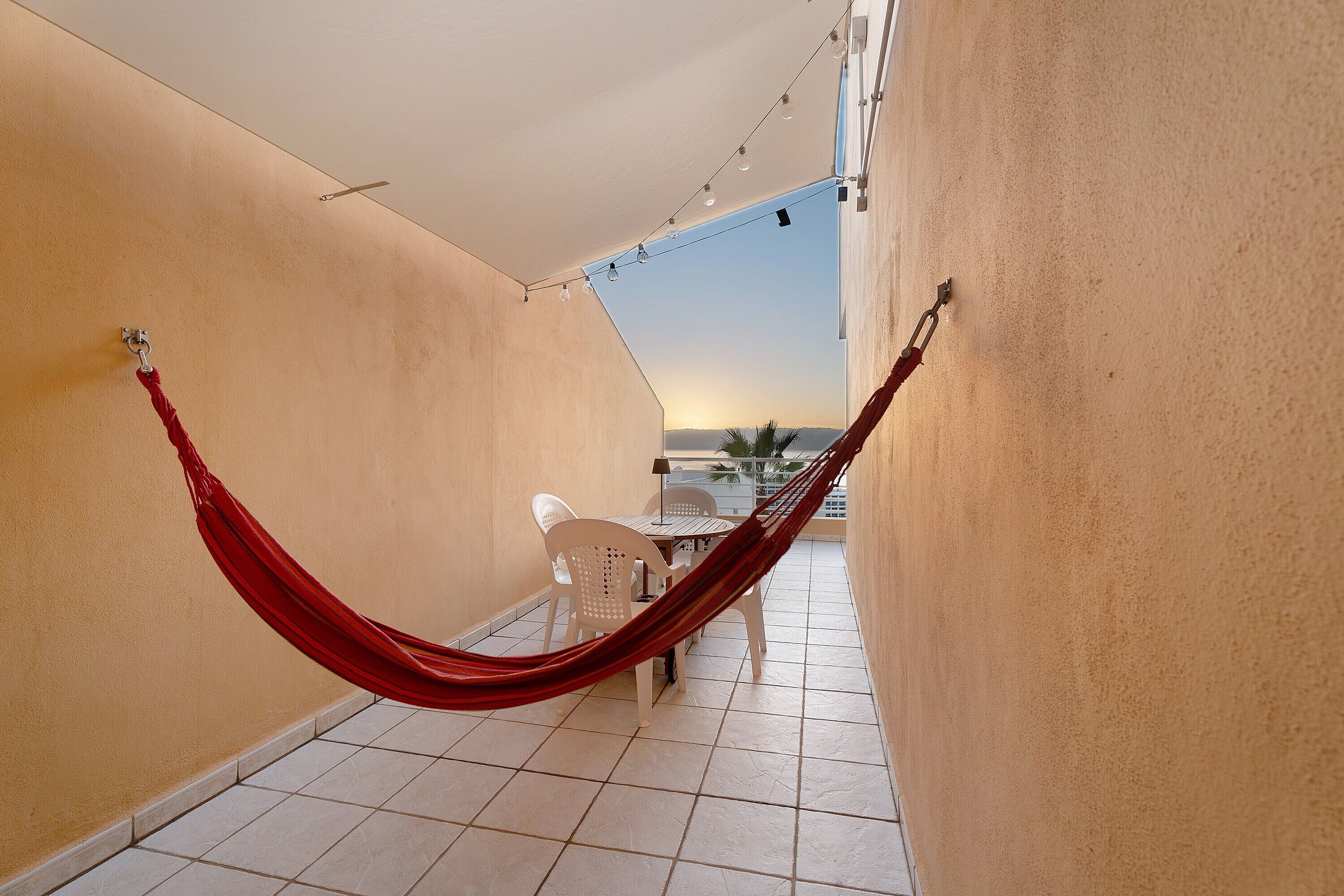 Un hamac sur un balcon, avec une table et des chaises, une vue sur le coucher de soleil et un sol carrelé.