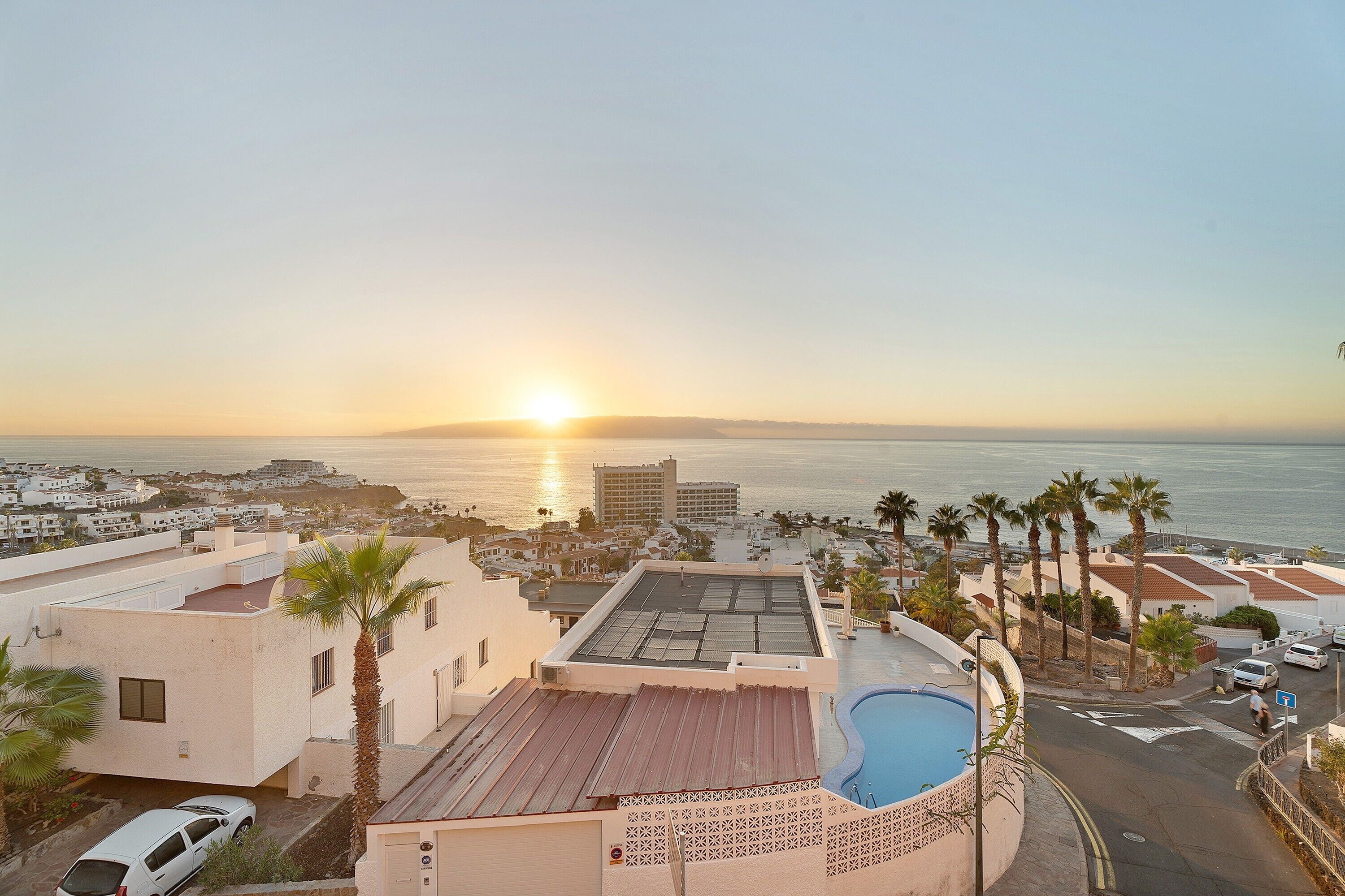 Un hotel frente al mar con piscina en la azotea, palmeras y vistas al atardecer.