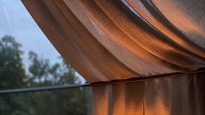 Una cama con un cojín estampado con motivos florales y una vela encendida en un recipiente de vidrio, frente a cortinas y una ventana con vistas a follaje.