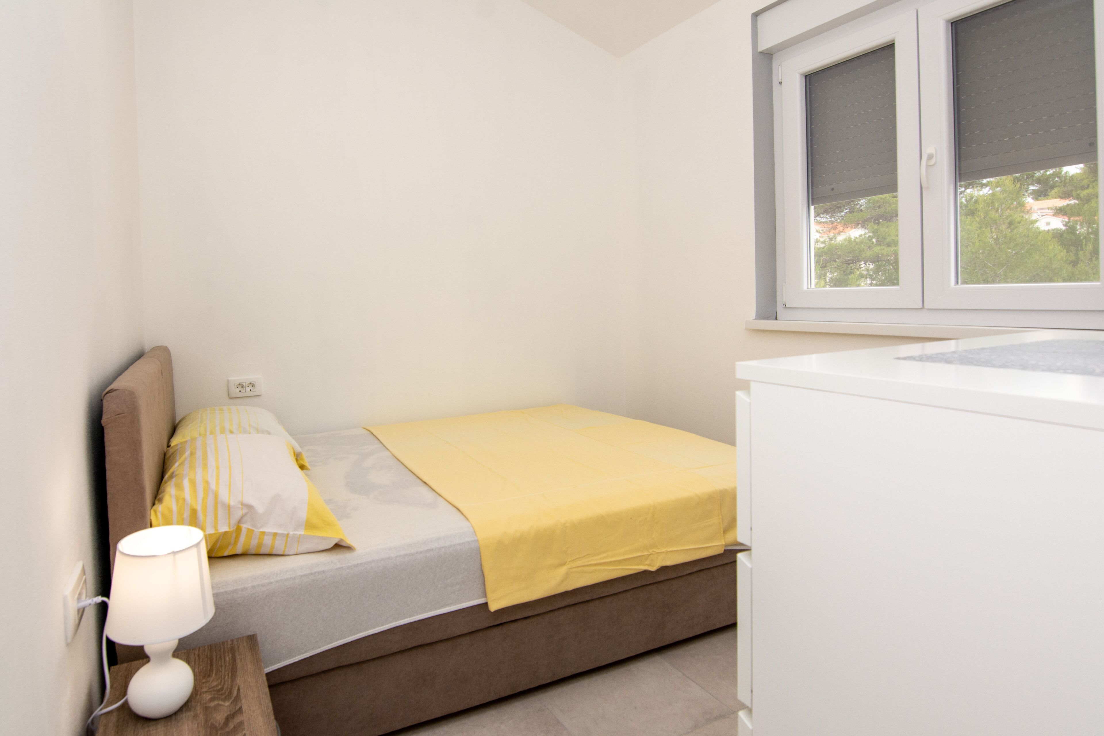 Un dormitorio pequeño y ordenado, con cama, lámpara de noche y ventana con persianas.