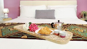 Un lit bien fait, avec du linge de lit blanc et un oreiller à rayures, un plateau avec des croissants, des fleurs et des tasses, et une table de chevet avec une lampe et des livres.