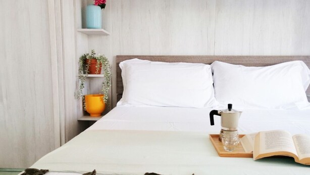 Un lit bien fait, avec du linge de lit blanc, une cafetière et un livre ouvert sur une table de chevet.