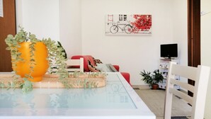 Un salon moderne avec une table à plateau en verre, un grand vase orange contenant des plantes vertes, un canapé rouge, un fauteuil blanc et un téléviseur posé sur un meuble blanc.