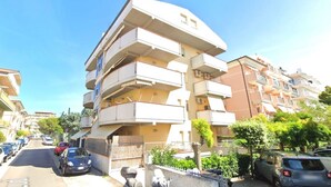 Un immeuble résidentiel à plusieurs étages, doté de balcons, avec des voitures garées et un ciel dégagé.