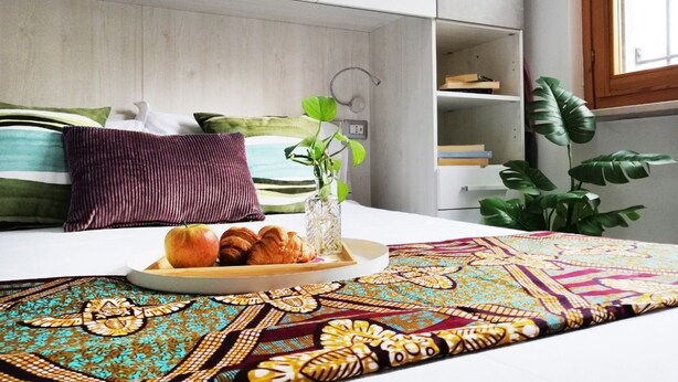 A neatly made bed with a tray of croissants and an apple, a patterned throw, and a potted plant. - HoliHome_Nobil Villani (Tortoreto Lido)