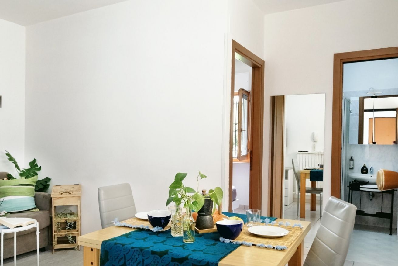 Uma sala de jantar com uma mesa de madeira, cadeiras e um jogo americano azul. Há plantas sobre a mesa e uma vista da cozinha ao fundo.