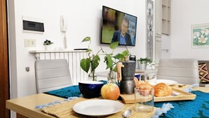 A dining area with a table set for two, featuring a plate, a glass, a bowl, a knife, a spoon, a bottle, and a croissant. There is a television mounted on the wall displaying a program. - HoliHome_Nobil Villani (Tortoreto Lido)