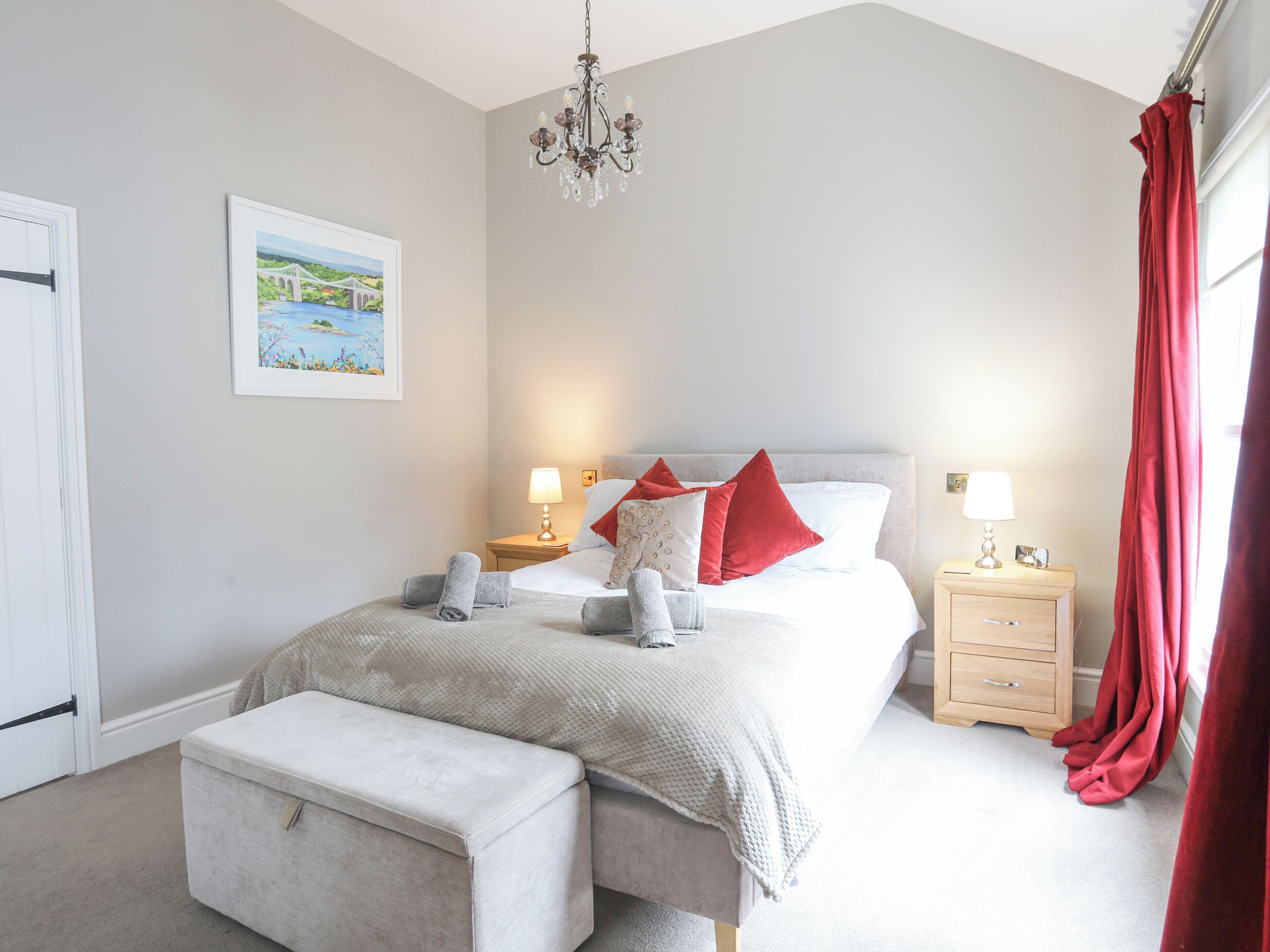 A bedroom with a bed, two bedside tables, a nightstand, a bench, a chandelier, and a window with curtains.