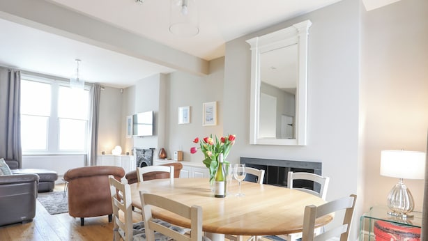 A spacious living room with a wooden dining table, white chairs, a sofa, and a fireplace. - Cobblers Cottage (Beaumaris)