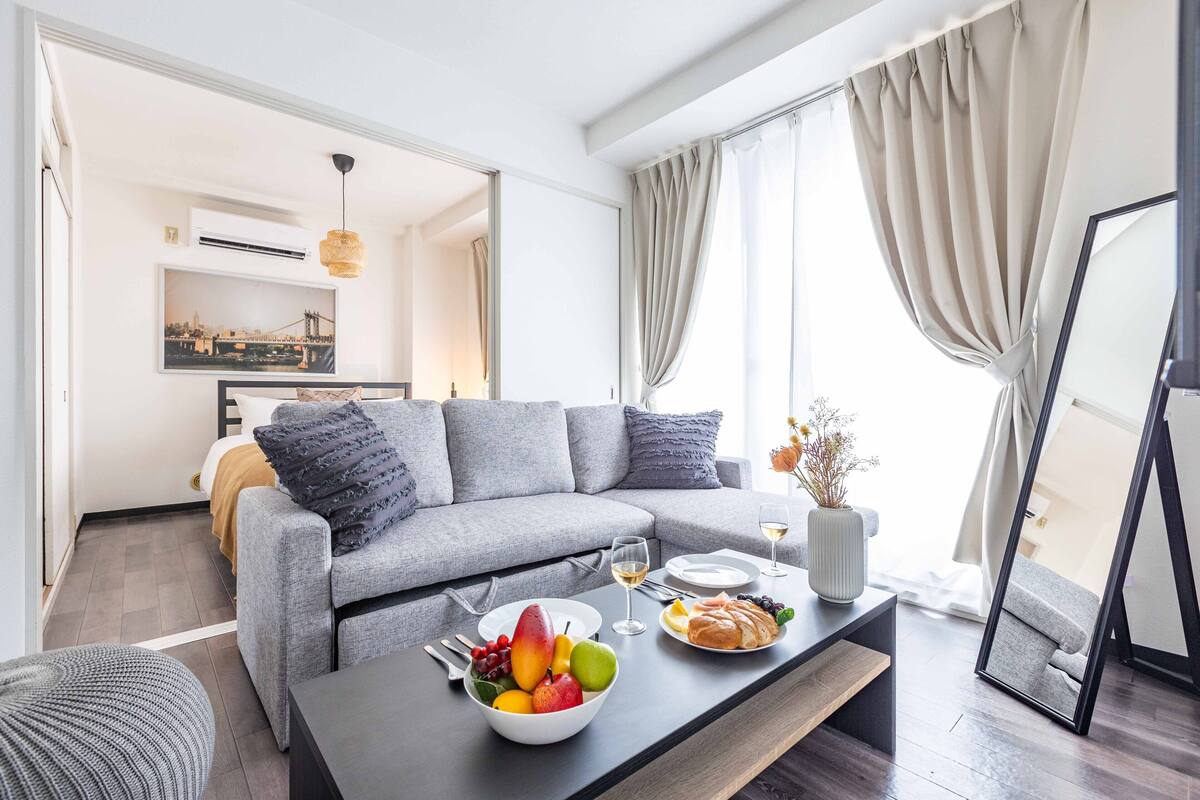 A modern living room with a grey sofa, a coffee table with fruit and pastries, and a dining area with a white table and chairs.