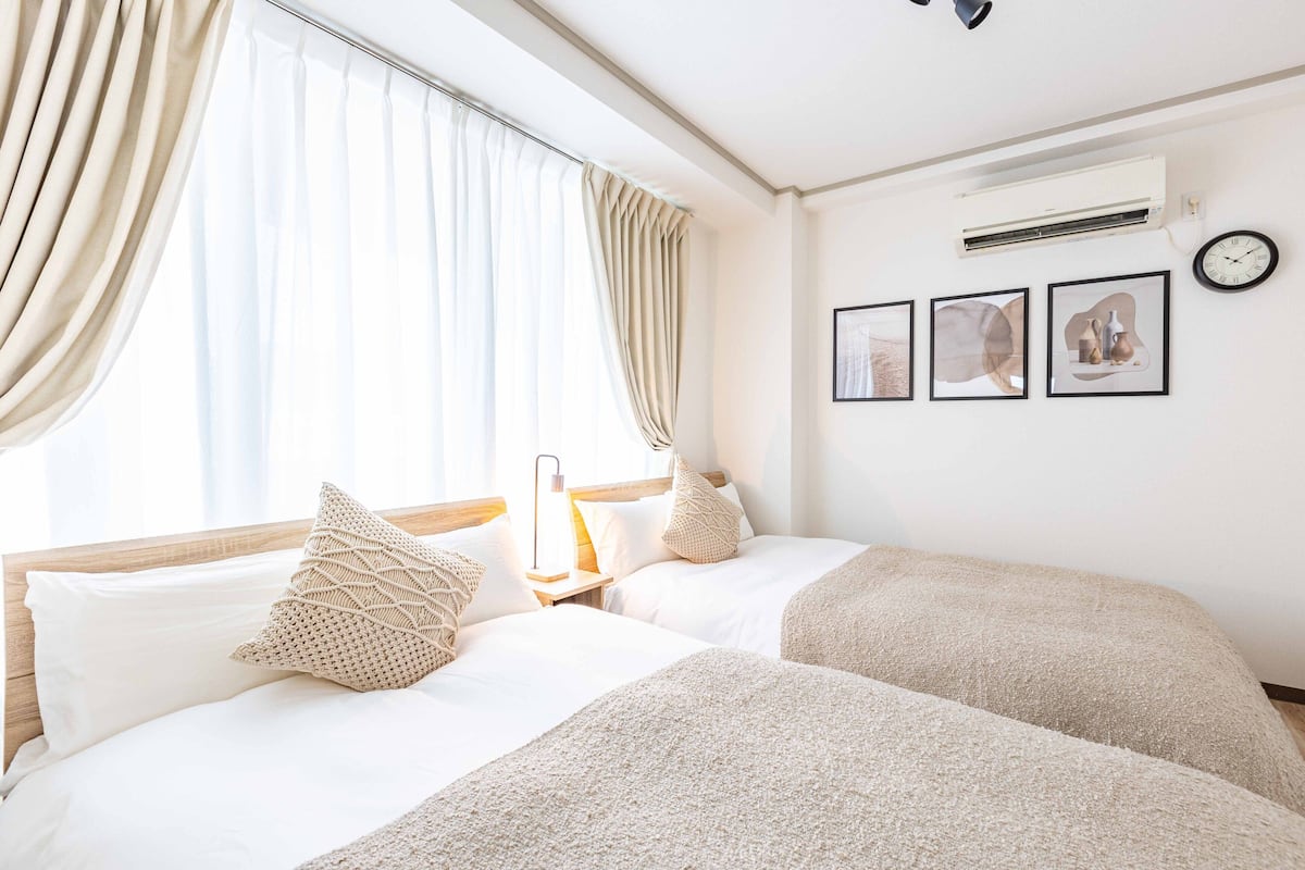A hotel room with two beds, beige curtains, a wall-mounted air conditioner, and three framed pictures.