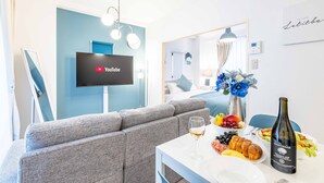A modern living room with a grey sofa, a flat-screen TV mounted on a blue wall, a dining table with a bottle of wine, and a fruit bowl. - STAY 1ST (Osaka)