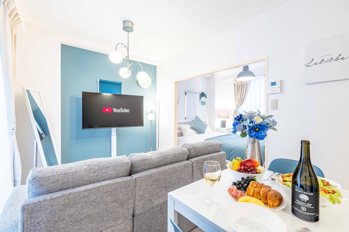 A modern living room with a grey sofa, a flat-screen TV mounted on a blue wall, a dining table with a bottle of wine, and a fruit bowl.