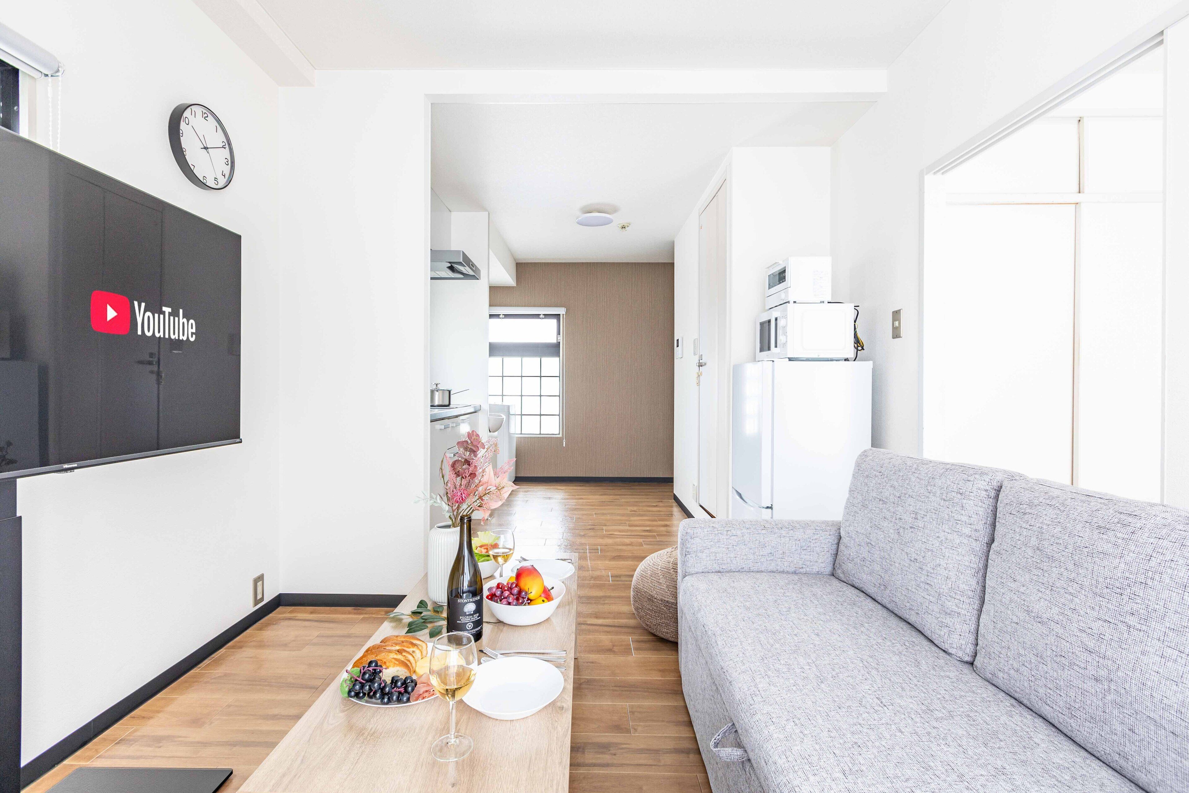 A modern living room with a grey sofa, a wooden dining table set with plates, glasses, and a vase of flowers. A flat-screen TV mounted on the wall displays the YouTube logo.