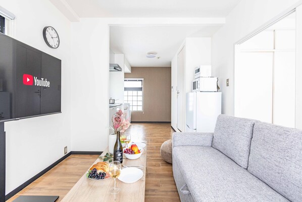 A modern living room with a grey sofa, a wooden dining table set with plates, glasses, and a vase of flowers. A flat-screen TV mounted on the wall displays the YouTube logo.