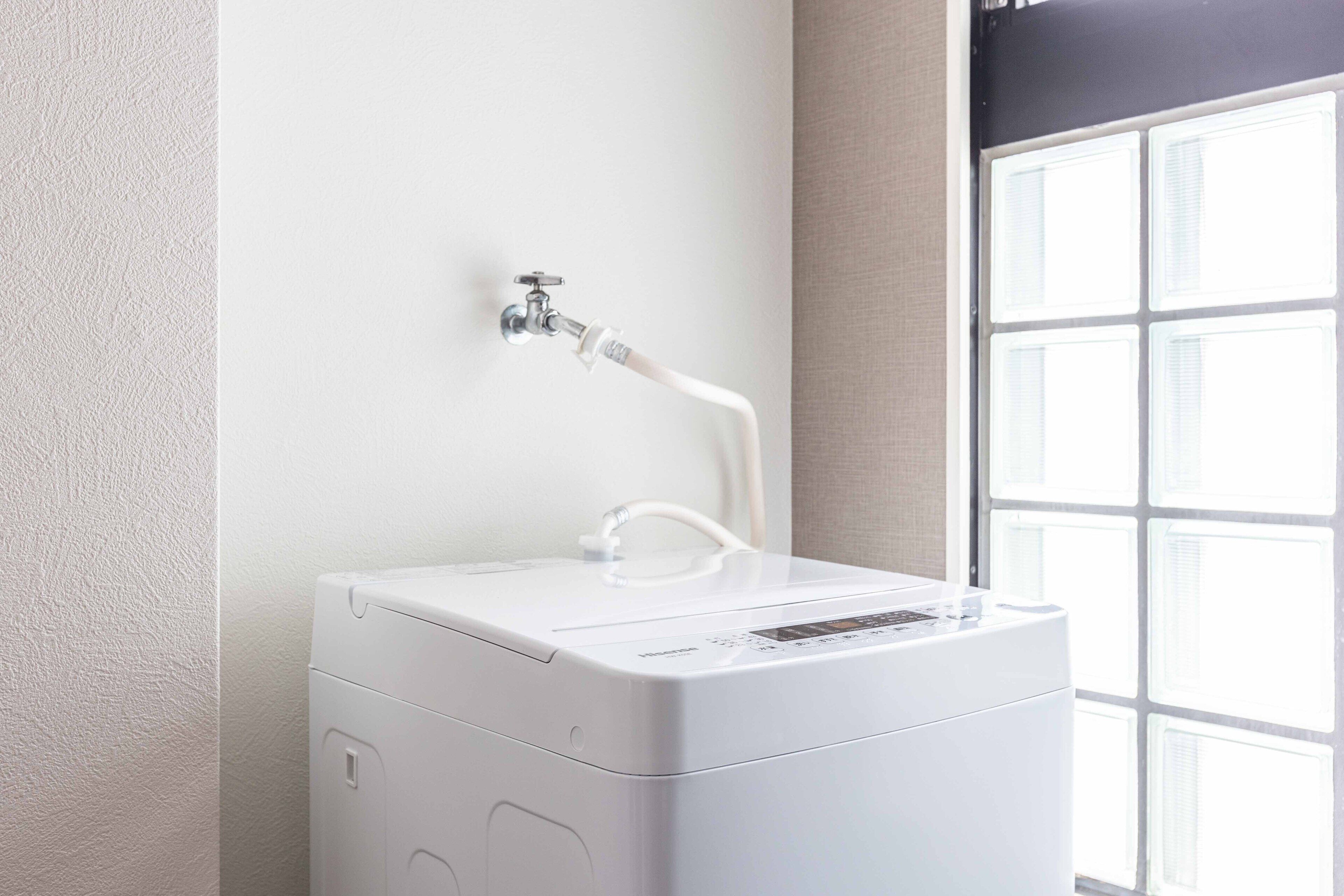 A compact, white, wall-mounted water heater with a hose and control panel, positioned in a room with a large window and a textured wall.
