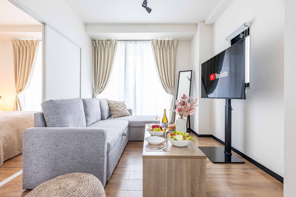A modern living room with a grey sofa, a wooden coffee table with a bowl of fruit, a bottle of wine, and a flat-screen TV mounted on the wall.