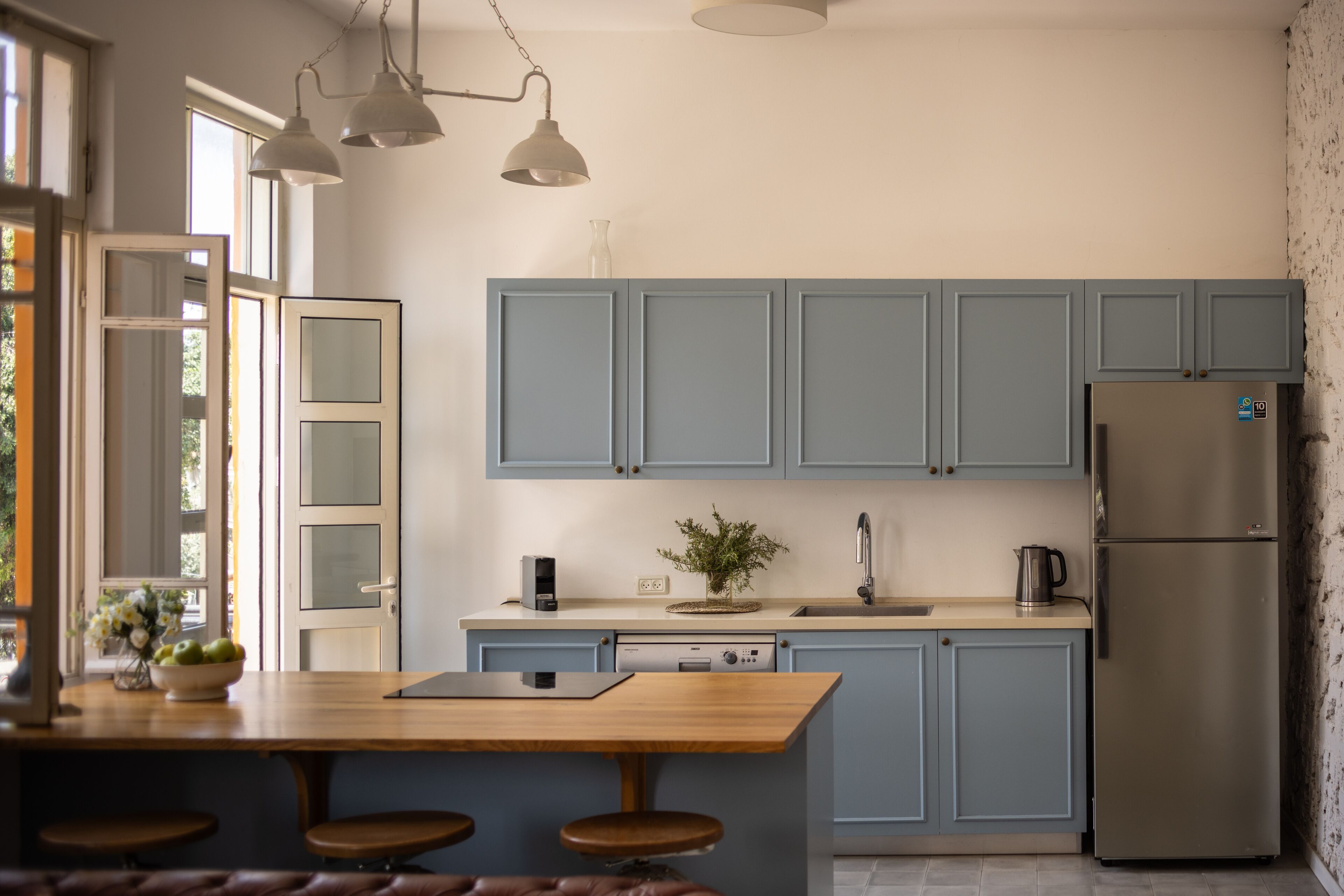 A modern kitchen with light blue cabinets, a stainless steel refrigerator, a wooden island with stools, and pendant lighting.