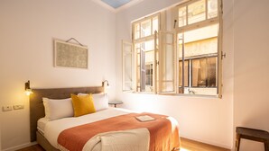 Ein Schlafzimmer mit einem Bett, einem Fenster mit geöffneten Lamellen, einem Nachttisch und einem Wandbild.