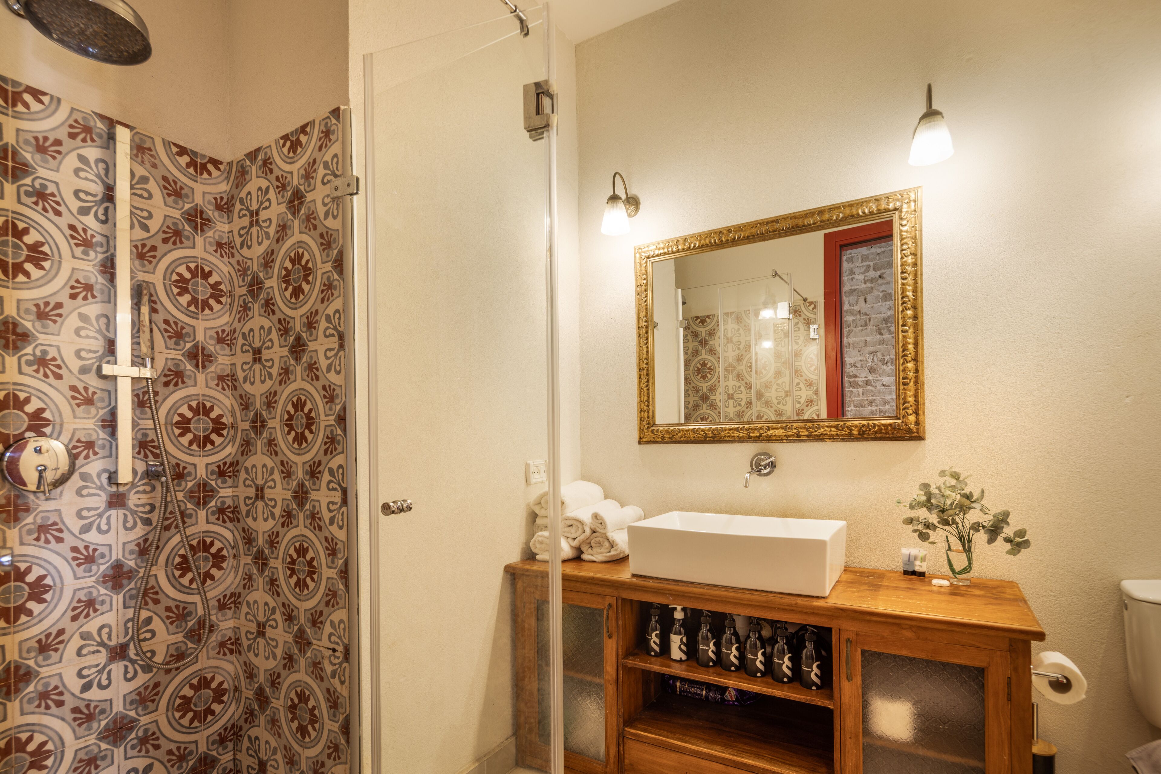 A bathroom with patterned tiles, a white sink, a mirror with a gold frame, and a wooden cabinet below the mirror.