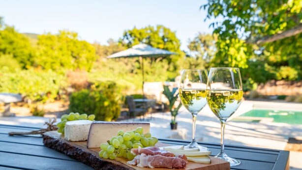 Una tabla de madera con queso, uvas y prosciutto sobre una mesa al aire libre, con una piscina y follaje en el fondo.