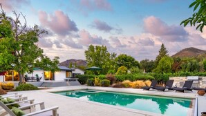 Una piscina con sillas de descanso, zona de barbacoa y una casa con mobiliario exterior.